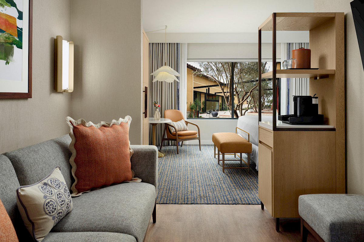 A modern hotel suite with a cozy living area, mid-century sofa and cushions in the foreground, a wooden shelf unit, and a view into a bright bedroom with a chair, ottoman, and large windows. The space opens to an outdoor courtyard through glass doors.